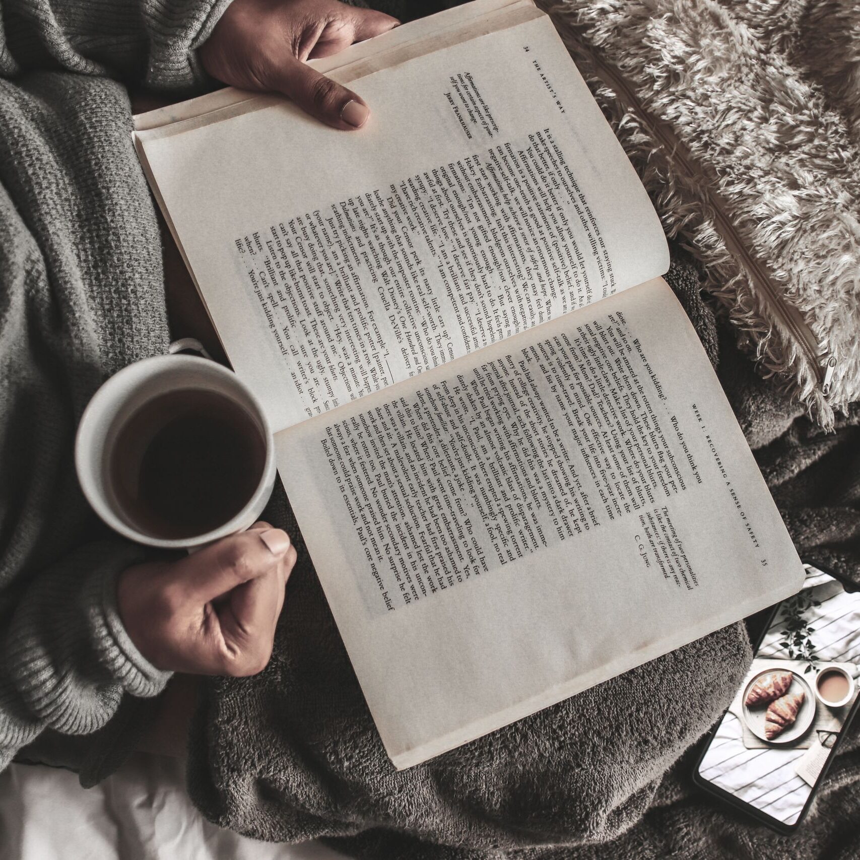 From above of crop anonymous female with mug of hot tea reading book under plaid near smartphone at home